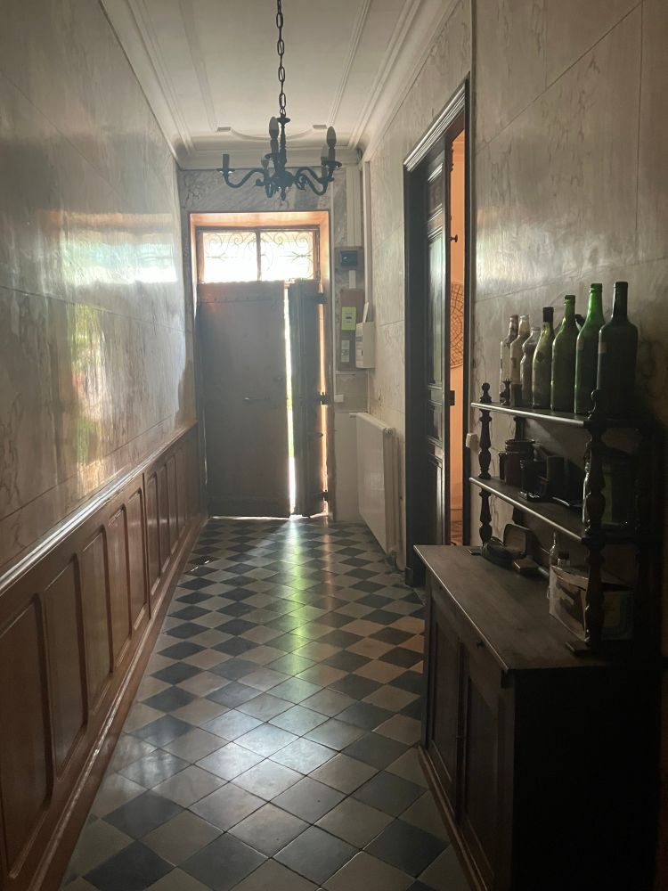 Couloir tout en longueur, carrelage noir et blanc en damier. Lourde porte en bois. À droit un meuble bas en bois et des bouteilles vides anciennes. Le mur est recouvert de bois sur la partie basse à gauche sur une hauteur de environ 1 mètre