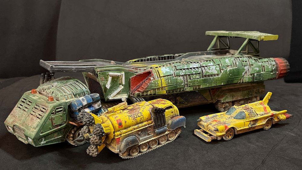 A photograph of the International Wreck Crew Gaslands team. Consisting of three minatures. The largest, Terrorbird Two, is a heavily converted car transporter with massive jet engines down each side, a large rear spoiler, and a futuristic raked cab at the front. Terrorbird Two is painted in green, with red and yellow accents, and a large white number two on the side of the trailer. This is clearly inspired by the iconic Thunderbird Two. 

In front of TB2 are the Mole and TB4. The Mole is tracked 1960s style small gas-tanker with an enormous tunnelling drill head at the front of the vehicle. The cab has been moved to the rear, and sits behind a set of massive exhausts. It is clearly inspired by the Mole from Thunderbirds, and is yellow with red accents and blue mudguards over the tracks. 

The third vehicle is Terrorbird 4. This is a version of Thunderbird 4 built from a Hot Wheels Adam West era Batmobile. It has added thrusters at the rear, and an array of tools and lights sticking out the front. The roof has been closed off with wire mesh and extra roof panelling. Like Thunderbird 4 she is bright yellow with red accents and a large number 4 on the side.

All the vehicles show significant wear, with rust, chipping, and weathering from desert sands.