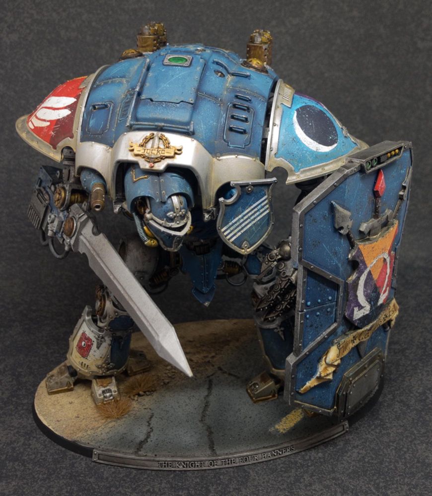 A photograph of a Warhammer 40,000 Imperial Knight miniature. The Knight has blue armour with silver trim, and is carrying a huge shield and sword.

This photo is front on. You can see the Knight’s armour including personal heraldry on the shoulders, and a huge silver sword-arm. 