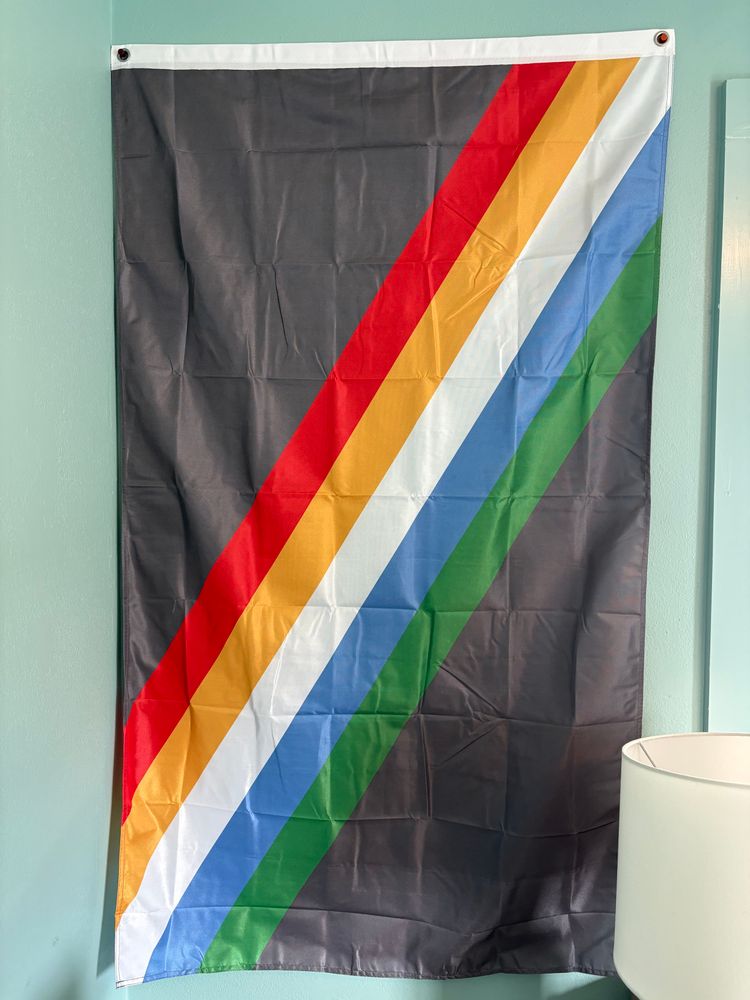Wrinkled disability pride flag hanging vertically with the red stripe on the left and two gromets on the top corners on a lightly saturated sea foam green wall with a lamp encroaching slightly on the bottom right. Window trim slightly visible on the right hand edge of the frame, not covering the flag.