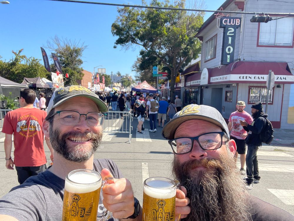 A selfie of Stephen and his twin brother holding frothy mugs of beer in front of an Oakland, CA street packed with people and vendor tents. 