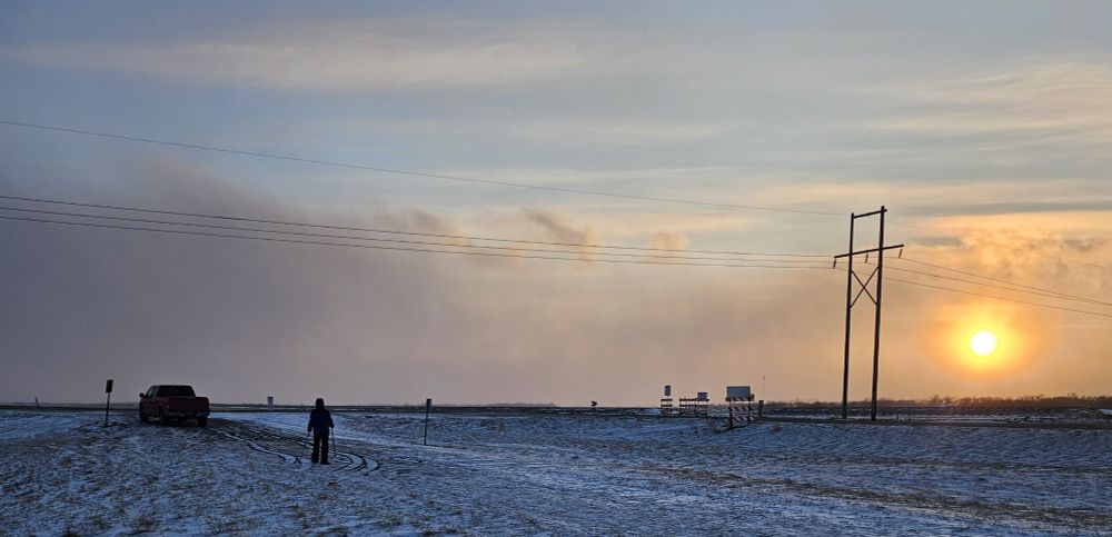A view of a boy with hiking poles and snowshoes nearing a pickup.  The background shows a setting sun obscured by some low, ice crystal clouds.