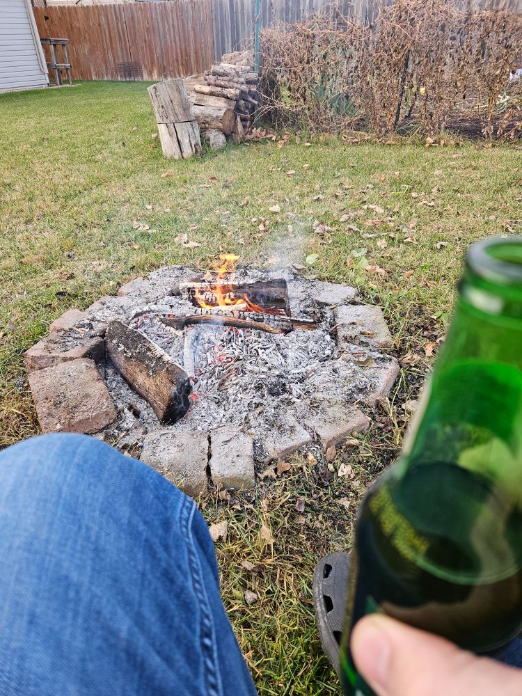 A knee in blue jeans and a beer in a green bottle are seen in the foreground.   A small fire burns in a fireplace.  Beyond is a pile of wood on the edge of a fenced garden.  On the fence hangs brown decaying vines.  In the background you see some wood fences.