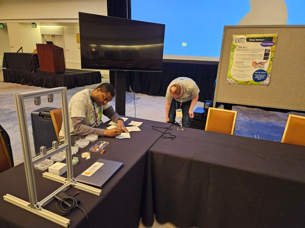 Leo-Stanley and Kurt are busy setting up an adhesion demo on some tables draped with long black tablecloths.