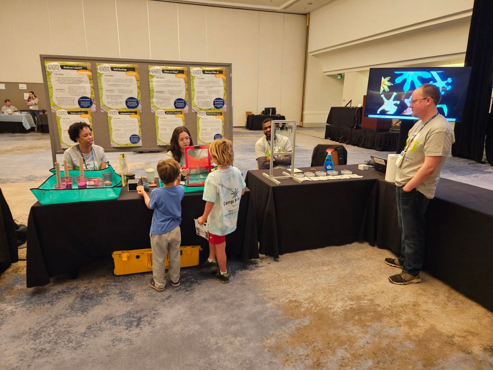Two children marvel at some granular physics!  Some red beads between two clear acrylic sheets show jamming as a scientist slowly opens a gap below the beads.  It is somewhat like a two dimensional hourglass.   Other scientists look on and many other demos sit on tables.