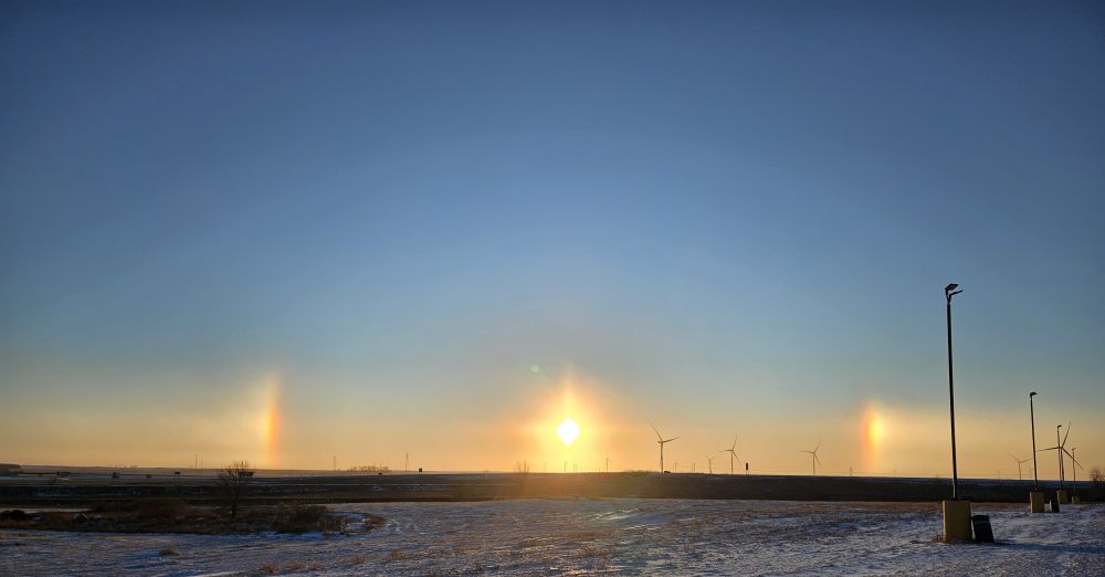 A photo of the snowy South Dakota prarie.  Center shows the sun near the horizon with two rainbow-like sun dogs on either side.  Turbines turn in the distance, while light polls in the right foreground slowly disappear into the distance.