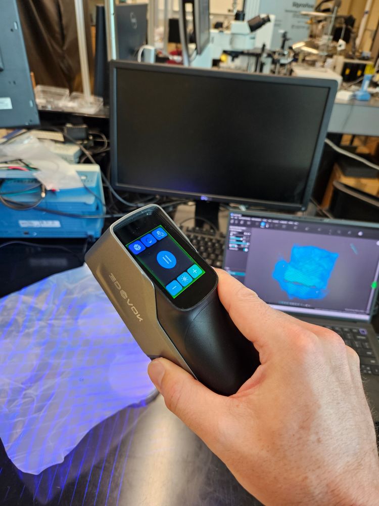 A hand holding a futuristic tool resembling a 1980's car phone in the foreground.   The device shines a grid of blue lasers onto a paper sheet.  Behind a computer screen shows the shape of the sheet.