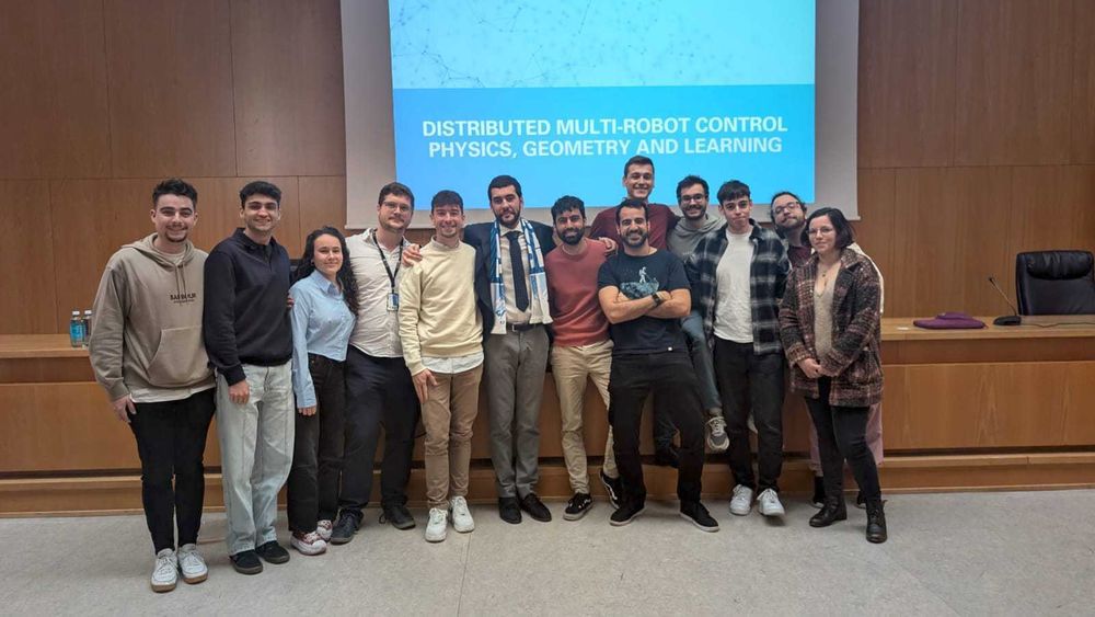Eduardo Sebastian, surrounded by other members of the RoPeRT group at a conference room posing happily in front of a title slide reading "DISTRIBUTED MULTI-ROBOT CONTROL, PHYSICS, GEOMETRY AND LEARNING"