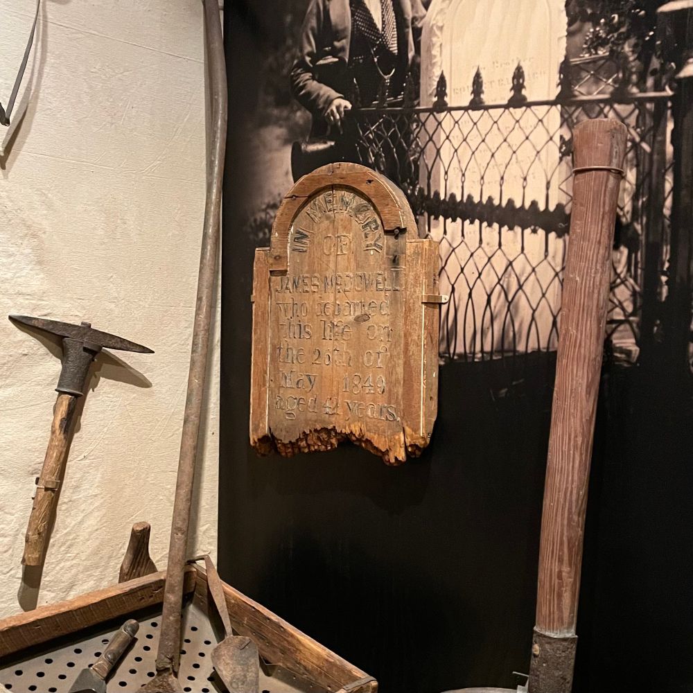 A wooden tombstone hanging on the wall in an exhibit that says: “In memory of James McDowell who departed this life on the 26th of May 1849 aged 42 years.”