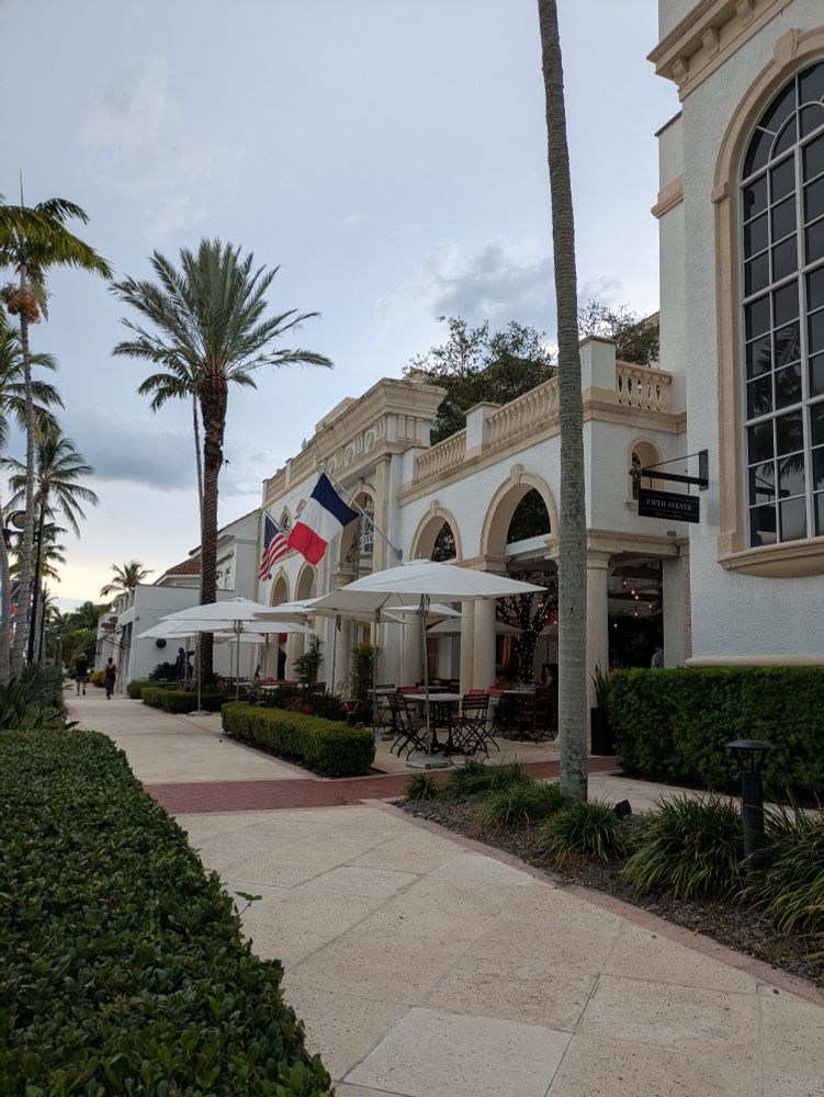 Building with arches and French bistro tables. There are palm trees out front and an American and French flag hung on the building. 