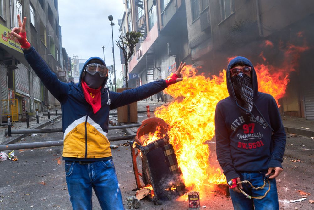 Das Bild zeigt zwei junge Personen, die vor einer brennenden Barrikade posieren. Beide tragen Gesichtsmasken und Schutzbrillen. Einer der beiden hebt die Hand zum Victory-Zeichen, während der andere eine entspannte Haltung einnimmt und eine Zwille in den Händen hält. Im Hintergrund brennt ein Möbelstück, und Flammen schlagen hoch. Die Straße ist unordentlich, mit Trümmern und anderen Gegenständen übersät.