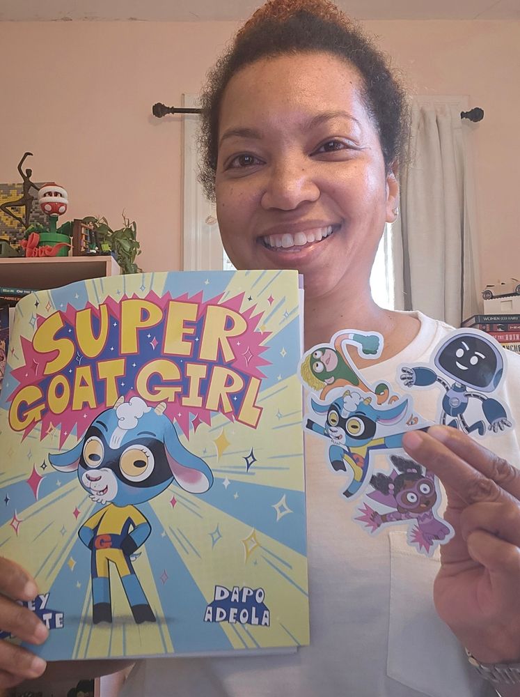 Smiling Black woman holding a copy of Super Goat Girl and a handful of stickers. 