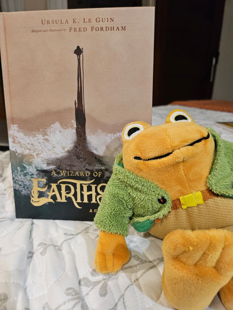 A photo of a plush Toad, from Frog and Toad are friends, sitting in front of the graphic novel version of A Wizard of Earthsea.