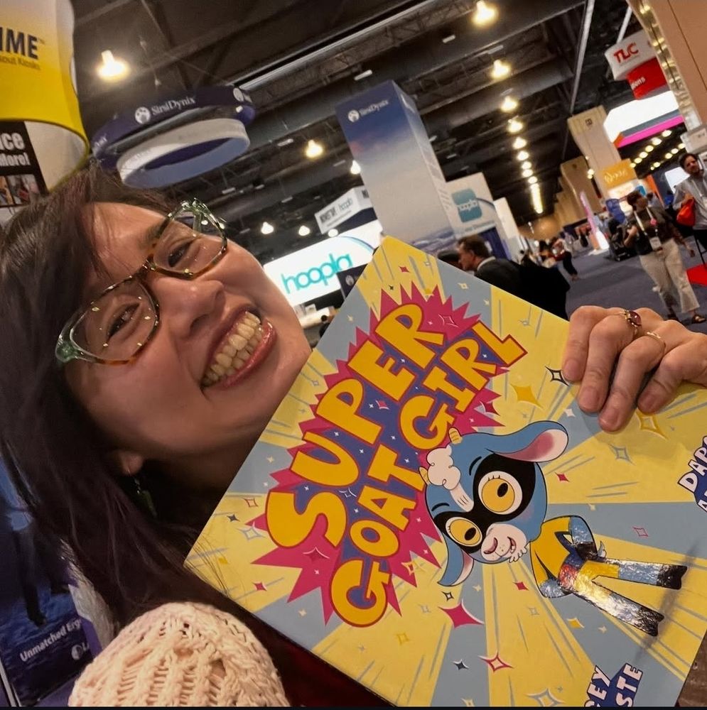 Smiling Asian woman wearing glasses holding a copy of Super Goat Girl. 