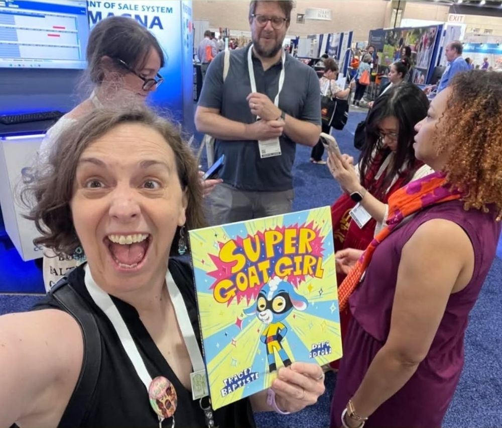 Grinning white woman holding a copy of Super Goat Girl in front of a group of people talking. 