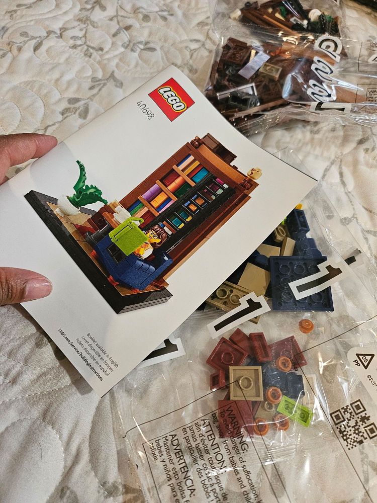 Photo of the instruction manual for the Lego set with the librarian and bookshelf. A couple of unopened bags of Legos are under the manual. 