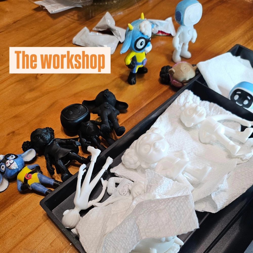 A photo labeled "The worksop" showing small figures in different stages of production. The figures are from the book Super Goat Girl. From left to right: Super Goat Girl fully painted with blue skin and yellow uniform, Laser Lass in all black resin and no paint, Robo Kid in all black work no paint, Super Goat Girl in all black with no paint, Super Goat Girl fully painted in shortalls, Robo Kid with lower body painted. 