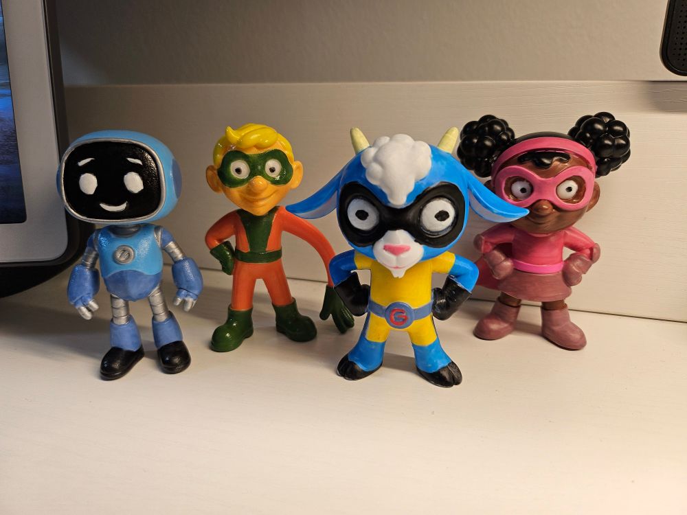 Four small figurines of characters from Super Goat Girl. Left to right: Robo Kid, a blue and silver figure with a screen face; Noodle Boy, an orange and green kid with blond hair and one overstreched arm; Super Goat Girl, a blue goat wearing yellow shortalls and a black eye mask; Laser Lass, a Black girl with afro puffs and an all-purple skirt ensemble including eye mask.