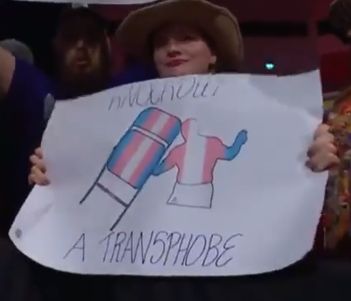 A woman (or someone presenting as femme) holding a sign in the crowd at the Utilita Arena in Cardiff, Wales. The sign features an outline of a wrestler holding a steel folding chair, both with the trans flag (blue, pink and white stripes) superimposed over them. The phrase "Knockout a transphobe" is written above and below.