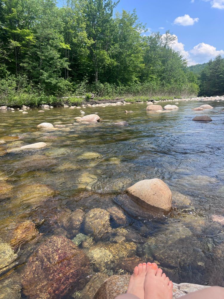 Pair of feet over a rock on a river side with forest in the background.