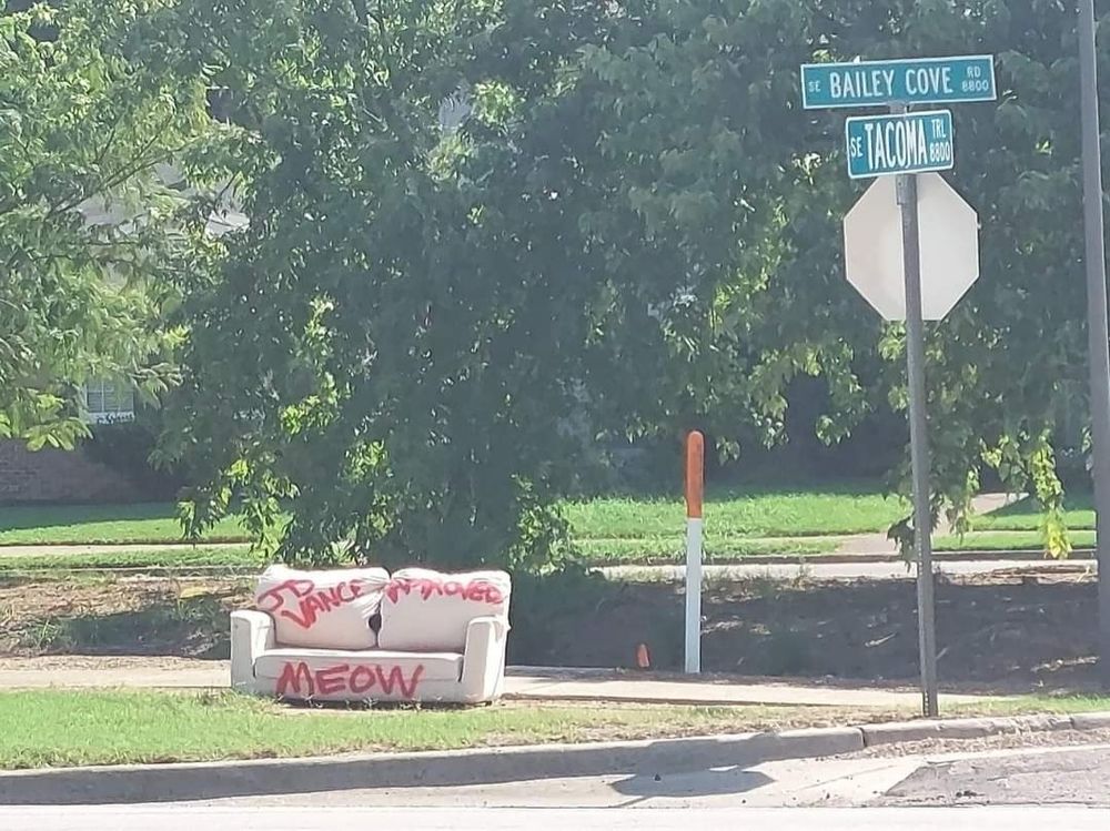 An abandoned white loveseat by the side of the road. Someone has spray-painted "JD Vance Approved" and "MEOW" in bright red paint on it.