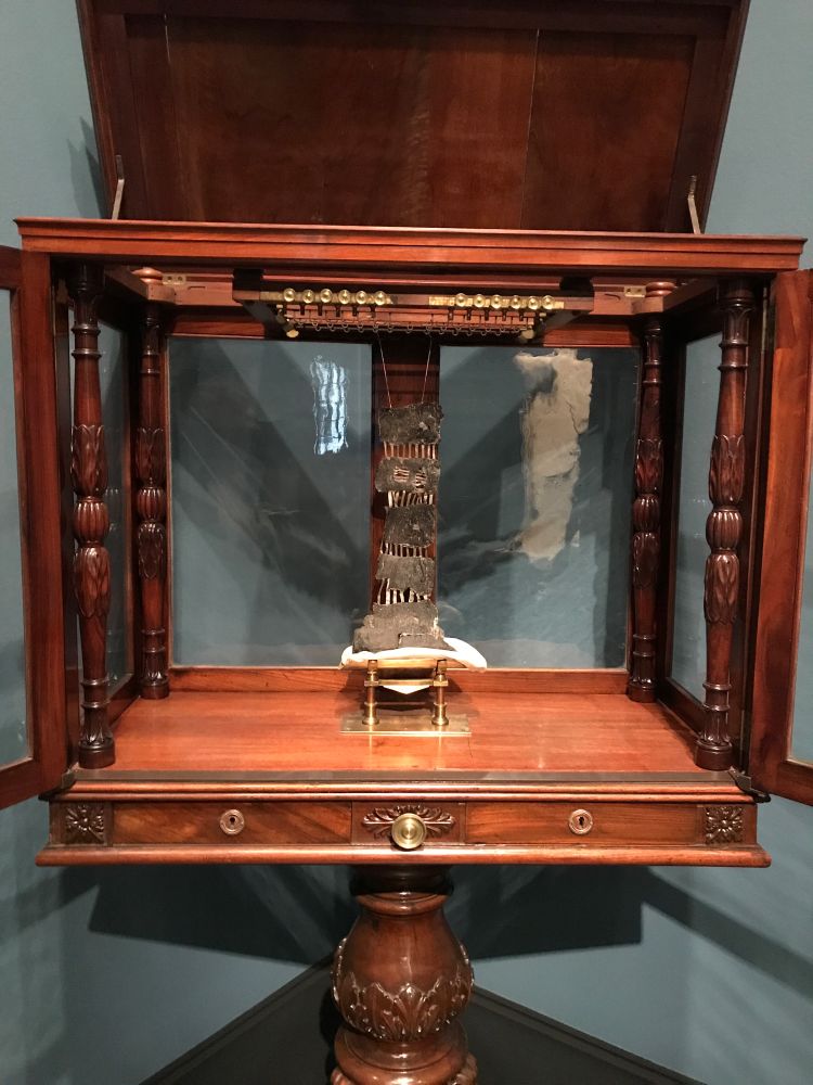 A fancy wooden cabinet with glass sides. At the centre, a strip of skeletal papyrus is partially unrolled, suspended from the ceiling of the cabinet.