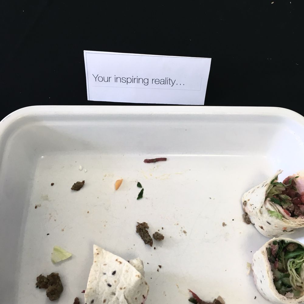 A partly empty tray of sandwich wraps, with crumbs. A small card behind it reads “your inspiring reality…”