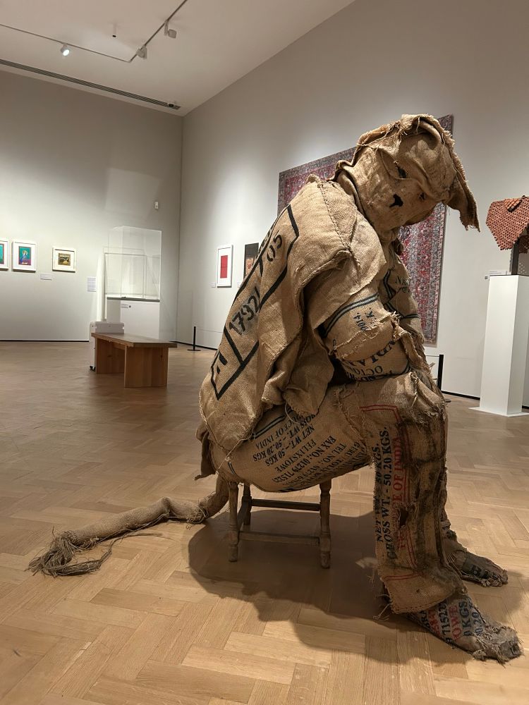 A figure made of coffee sacks rests on a tiny stool in a gallery. Part human, part big cat - a long tail curls behind and feline ears droop at the head. Faint details of a face suggest a cat-like nose. 