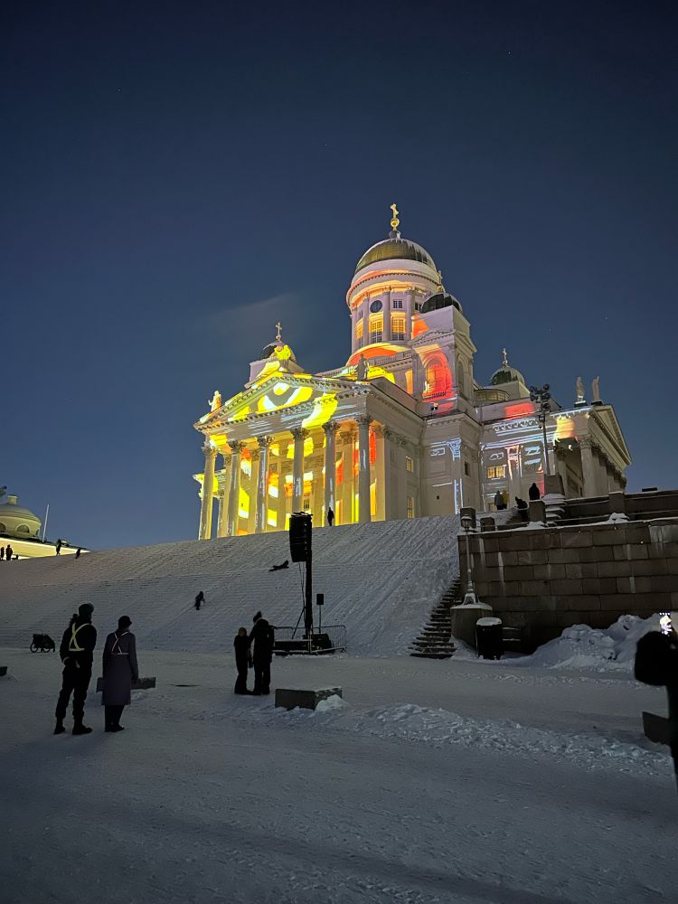 Keltaisilla ja punaisilla kuvioilla valaistu Helsingin Tuomiokirkko talvella, ihmisiä ihastelemassa valotaidetta.