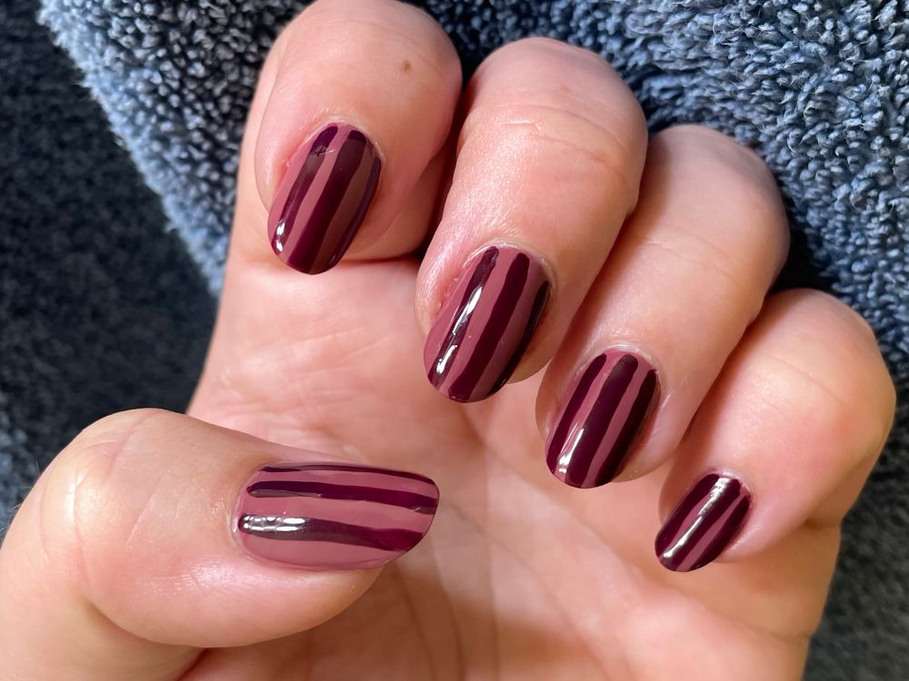Photo of a hand loosely curved to show its manicure. The fingernails are painted deep pink with purple vertical stripes. 