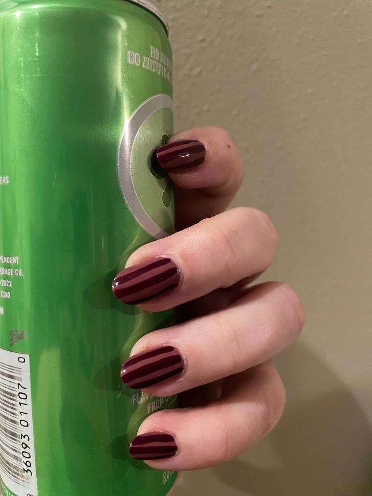 Photo of four fingers curved around a green can (the thumb is out of frame). The fingernails are painted deep pink with purple vertical stripes. 