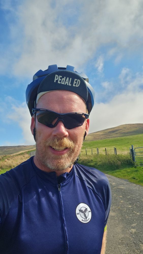 Michael in navy cycling jersey with green hills, blue skies and white clouds in the background