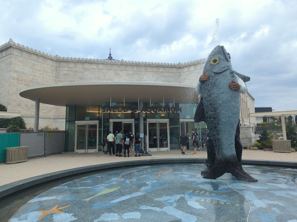 The entrance to Shedd Aquarium: a tan stone building with a fountain out front made from a person holding a fish