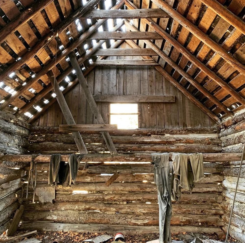 The inside of an old decrepit A-frame log home from the 1800s, with clothing and other remnants strewn about. there is a small bright window in the centre of the frame. 