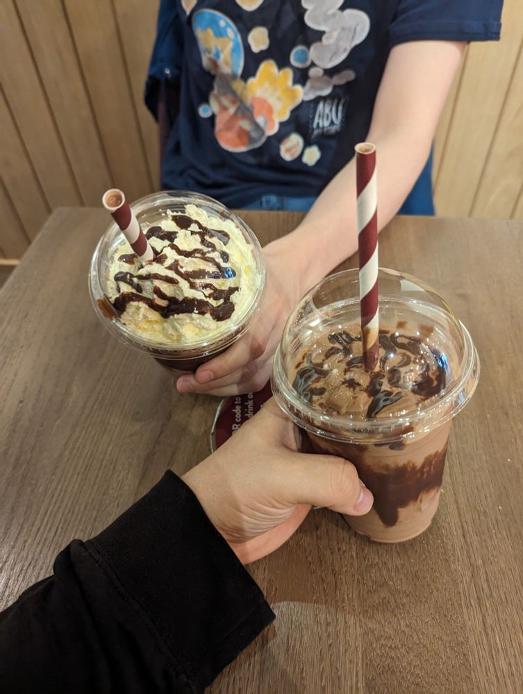 An image at a table in Costa Coffee, from my POV, with us both holding a Chocolate Brownie Frappe. Shifty's has cream on top, mine doesn't, but they're both drizzled with chocolate.