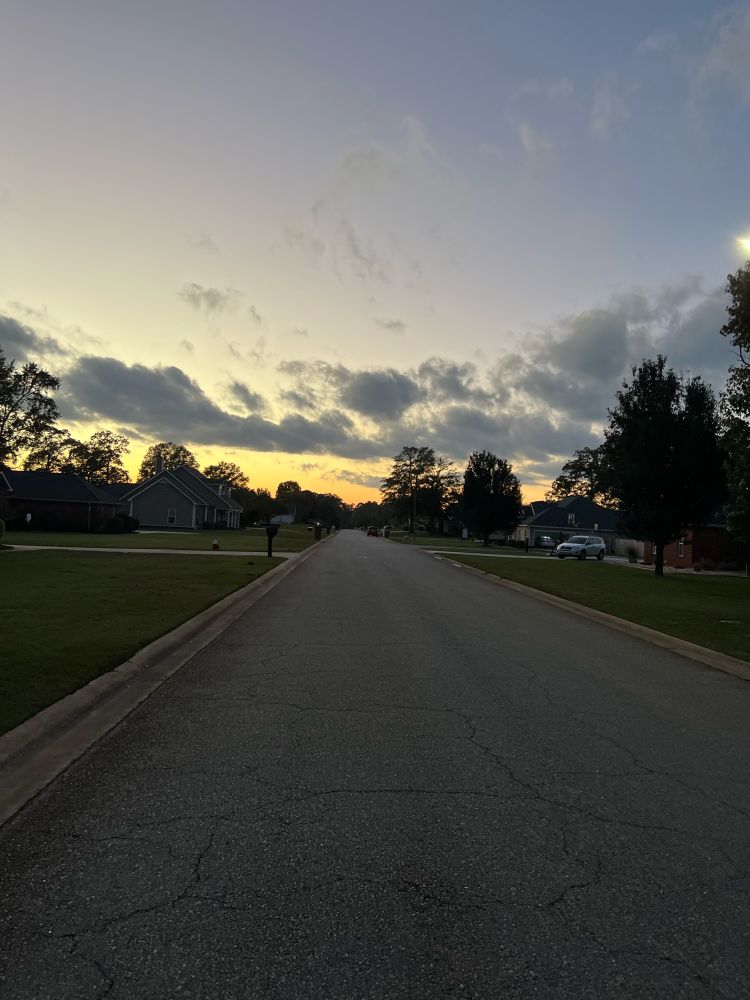 A lightly colored sunset at the end of a long residential neighborhood street. The street is flanked by brick homes and mailboxes with the occasional vehicle in a driveway. 
