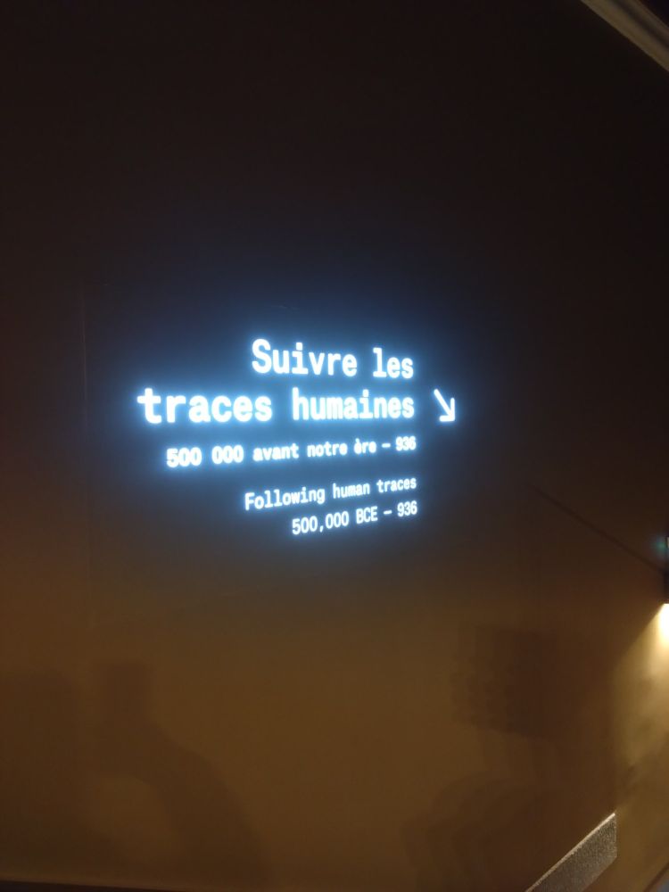 Inscription lumineuse sur un mur indiquant "suivre les traces humaines 500 000 avant notre ère - date peu lisible" et la traduction en anglais. Une flèche pointe vers le bas à droite.