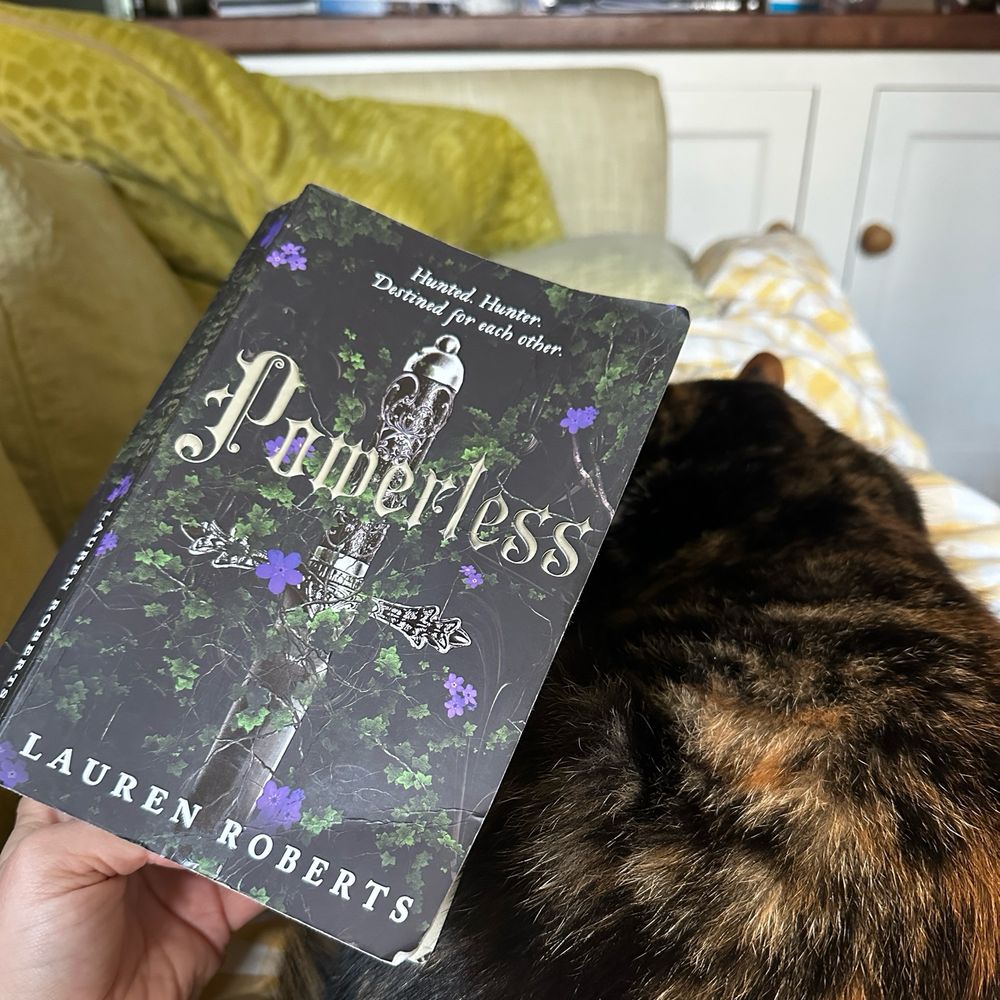 Powerless by Lauren Roberts in my left hand and a tortie cat on my lap to the right of it.