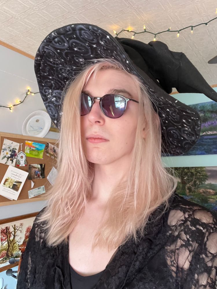 a selfie of ursula, fae is wearing a black witch hat, dark purple sunglasses, a lace top over a black bra. 