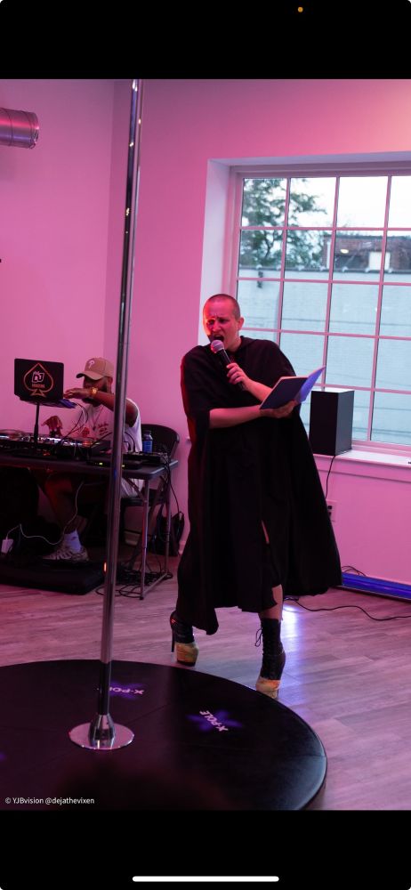 Rissa, adorned with a cape and platforms, holding a book in one hand and speaking into a microphone. There's a DJ behind her and a pole in front of her.