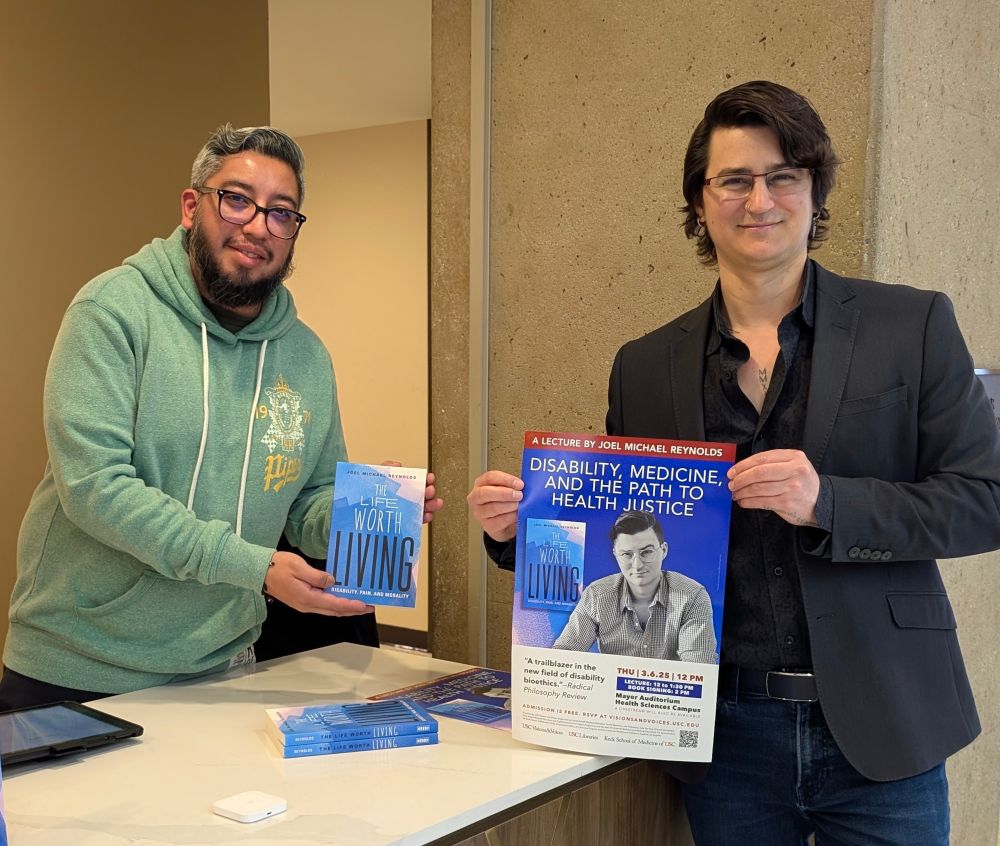 Joel Michael Reynolds (in a black button up and dark gray blazer) holding a poster for their talk "Disability, Medicine, and the Path to Health Justice" as part of the USC Visions and Voices 2025 series. They are standing next to an employee from the bookstore who is holding their book, "The Life Worth Living: Disability, Pain, and Morality."
