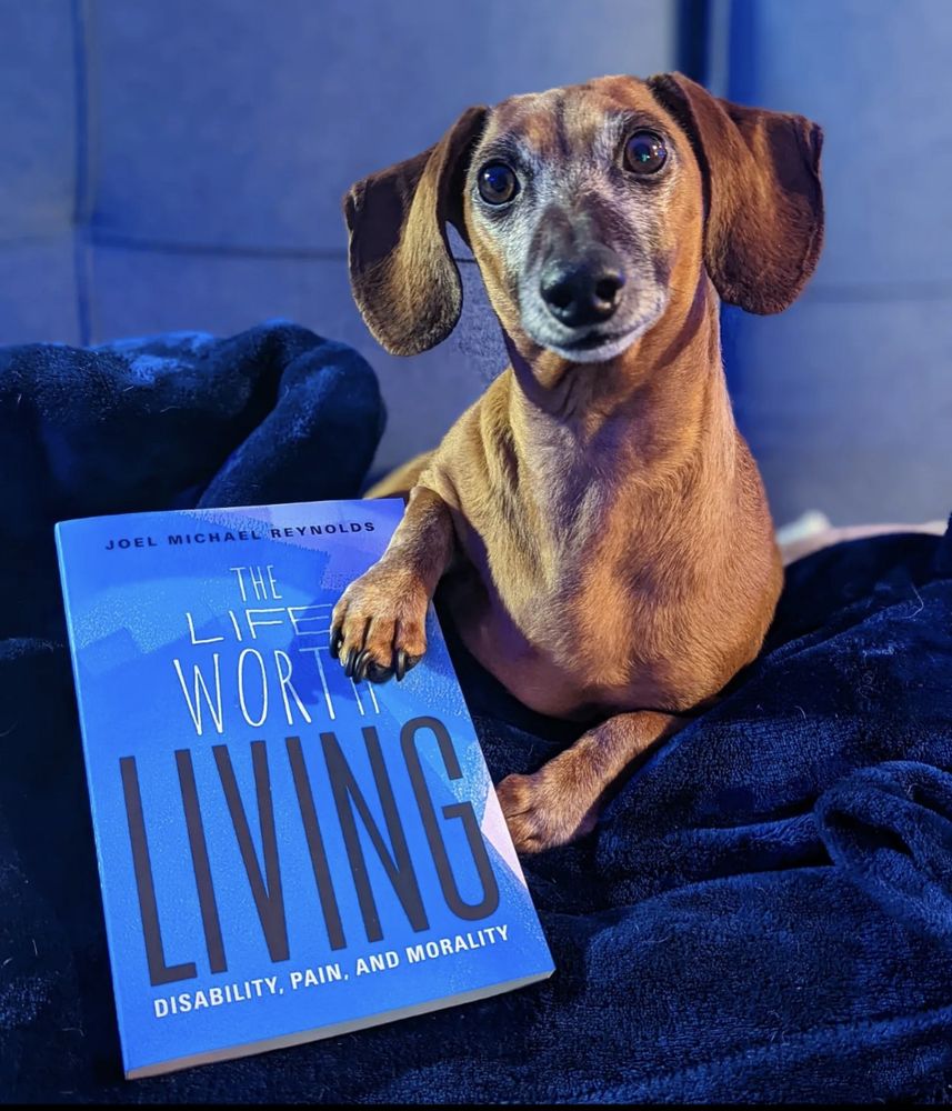 An adorable miniature dachshund looking directly into the camera with his front right paw on the book, "The Life Worth Living: Disability, Pain, and Morality" by Joel Michael Reynolds.