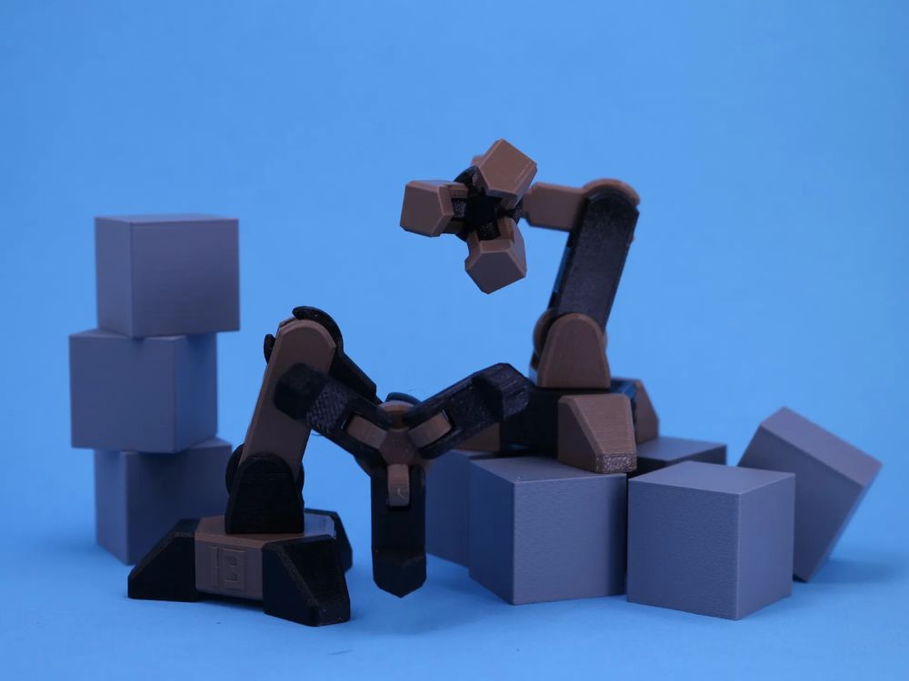 Two small 3D printed robot arms, posed as though looking at the camera with their claw "faces." They are black and light-brown, posed among gray cubes on a blue background.