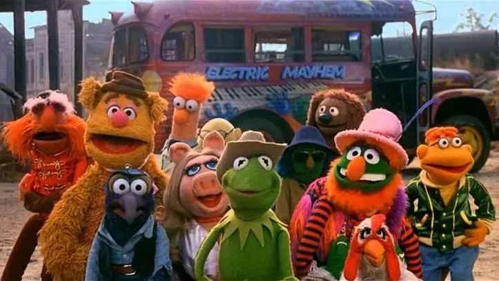 A still from the Muppet Movie with Kermit the Frog and a group of Muppets including Miss Piggy, Gonzo, Fozzie Bear and Dr. Teeth and the Electric Mayhem band. They are standing in front of the band tour bus, half way along on their show biz adventure. Kermit wears a cowboy hat because he is a bad-ass.