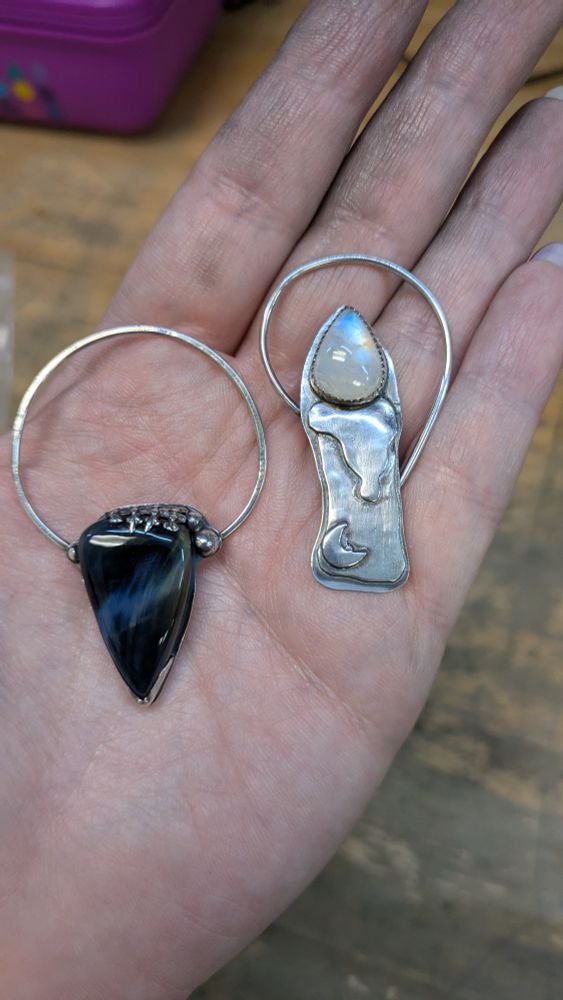A pale hand has two silver pendants. One is abstract with a partial asymmetrical solid bezel holding the bottom point and Gothic style pronged bezel holding the top of a Petersite, a black stone with a blue sheen. The other is a melting candle with a moon near the bottom and a rainbow moonstone set as the flame.
