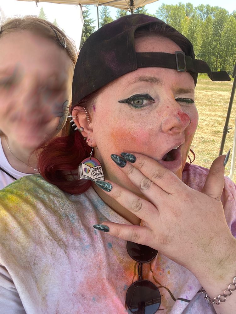 A photo of a woman covered in multicolored powder being photobombed by a teen. The teens face is blurred. 