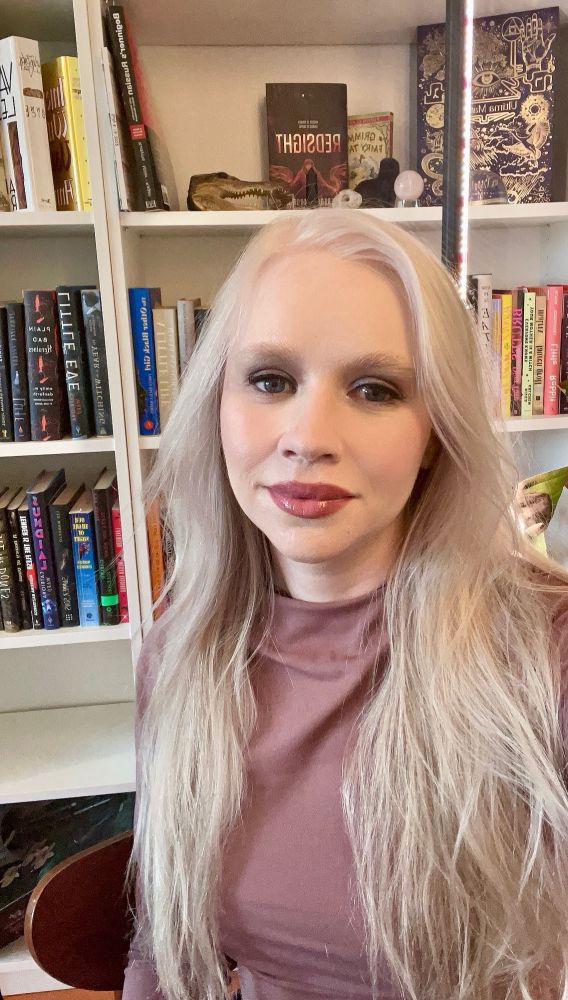 a photo of me wearing a pink shirt sitting in front of a bookshelf with my hair down.