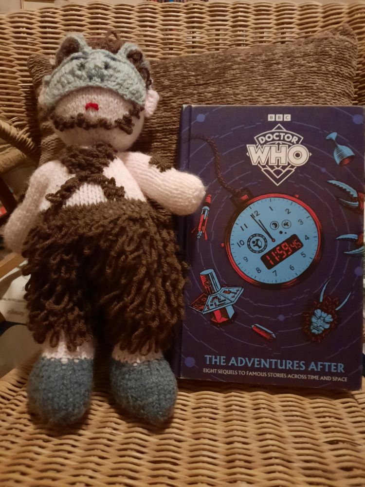 A knitted character with woolly hairy legs, a bare chest and a horned mask is standing next to a book with a blue cover. The book is entitled "Doctor Who, the adventures after"