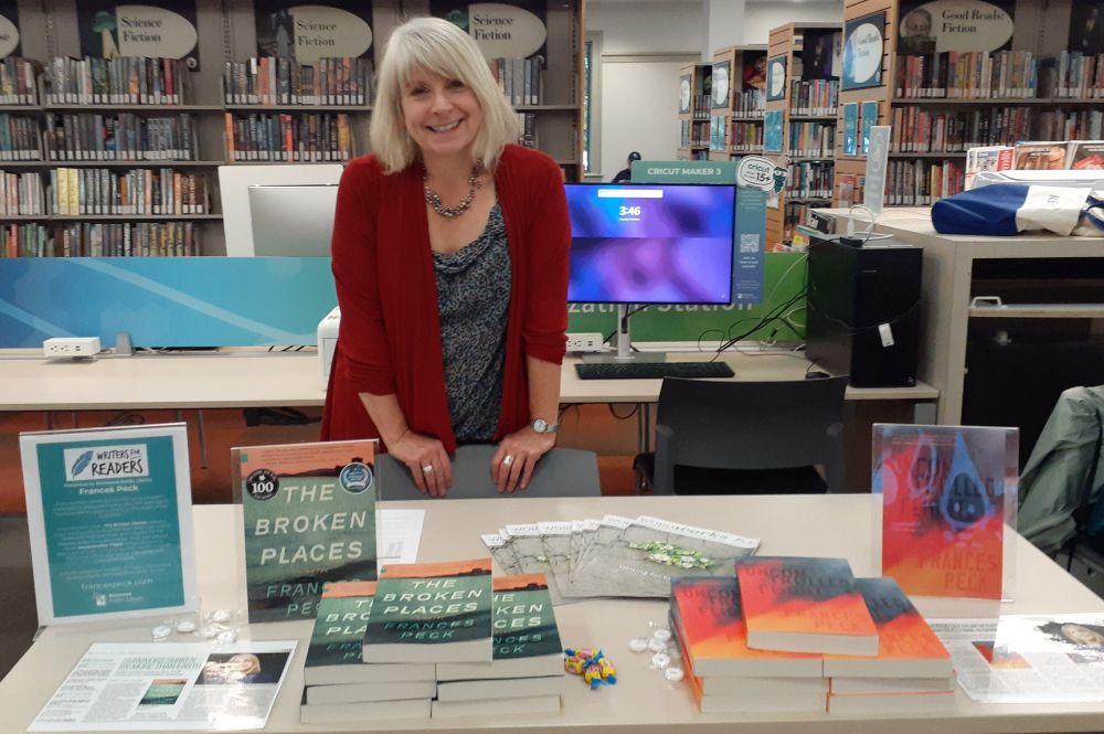 Blond white woman in a red sweater standing behind a table of books, with library bookcases in the background.