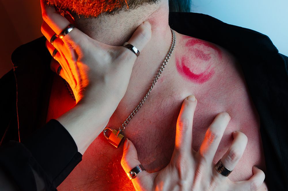 Nahaufnahme eines nackten Oberkörpers, teilweise bedeckt von einem offenen schwarzen Hemd. Eine Kette mit einem kleinen Vorhängeschloss hängt um den Hals. Auf der Haut ist ein deutlicher Lippenstiftabdruck zu sehen, in dessen Mitte sich ein Saughämatom befindet. Zwei Hände einer anderen Person mit mehreren Ringen greifen den Hals und die Brust. Eine davon umschließt leicht die Kehle unterhalb des Bartes. Die zweite Hand krallt sich in die Brust. Das Licht ist eine Mischung aus kühlem Blau und warmem Orange, das einen starken Kontrast erzeugt.